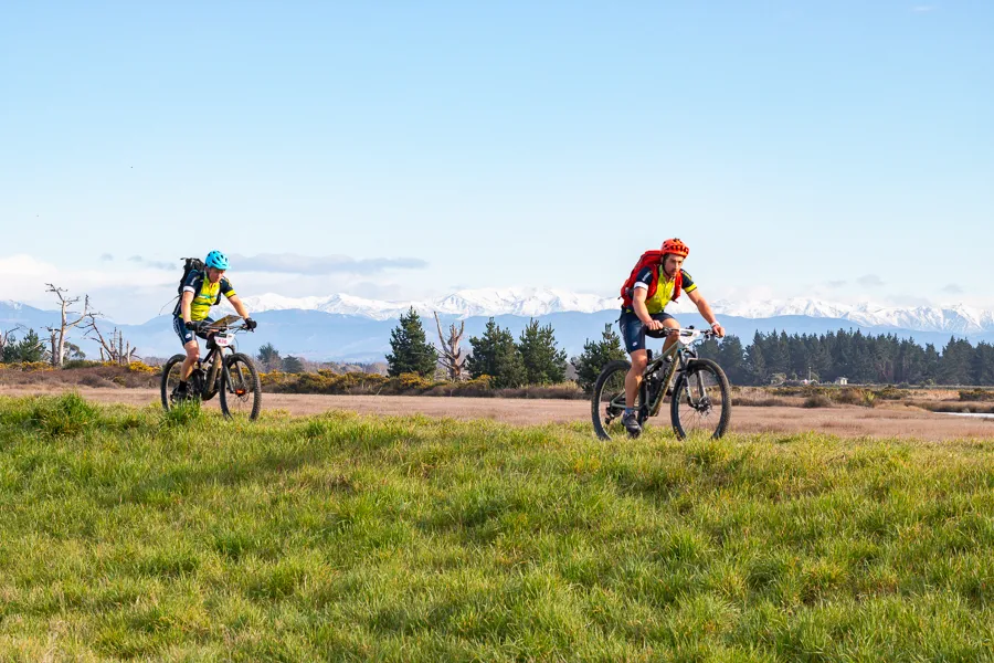 Two riders cross open farmland with the Southern Alps behind