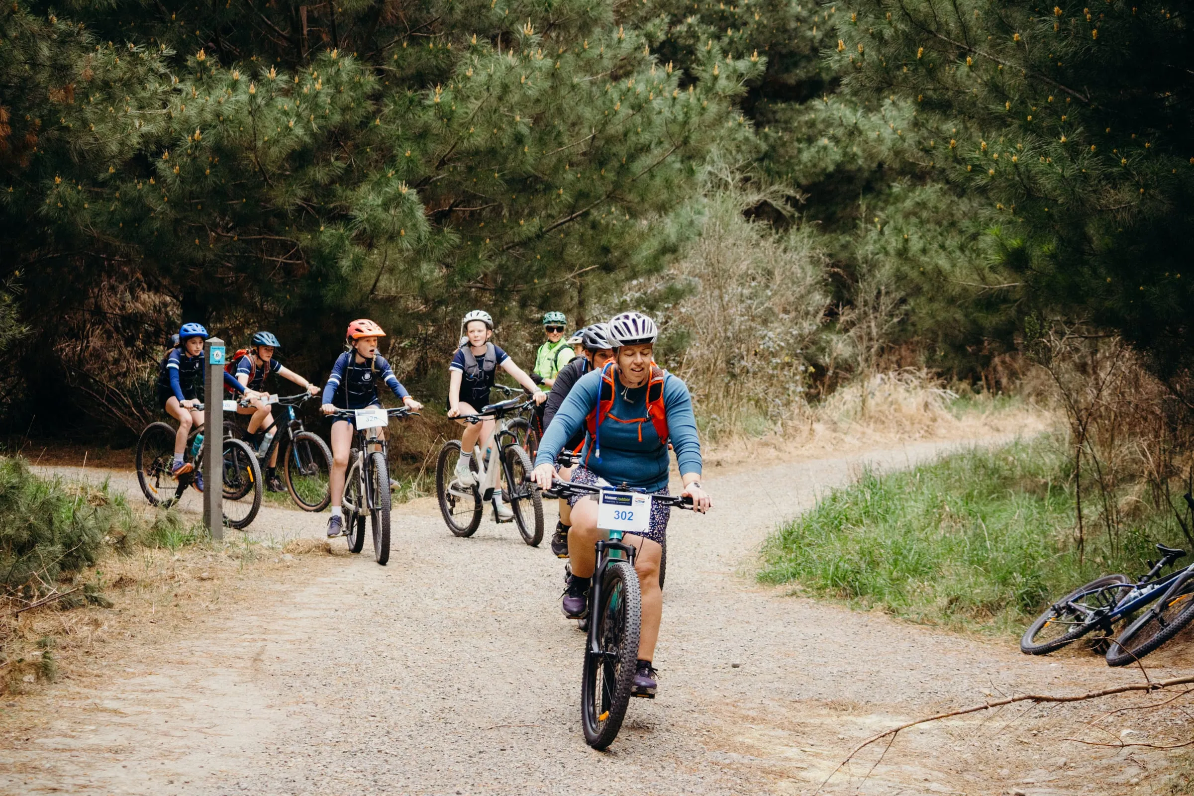 A line of riders on a forest gravel track, a woman in front with her number board on the bars
