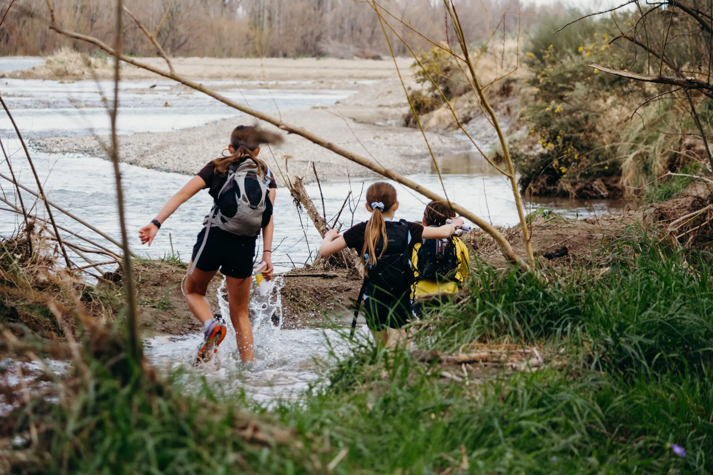 Three racers splash across a braided river channel with their packs on